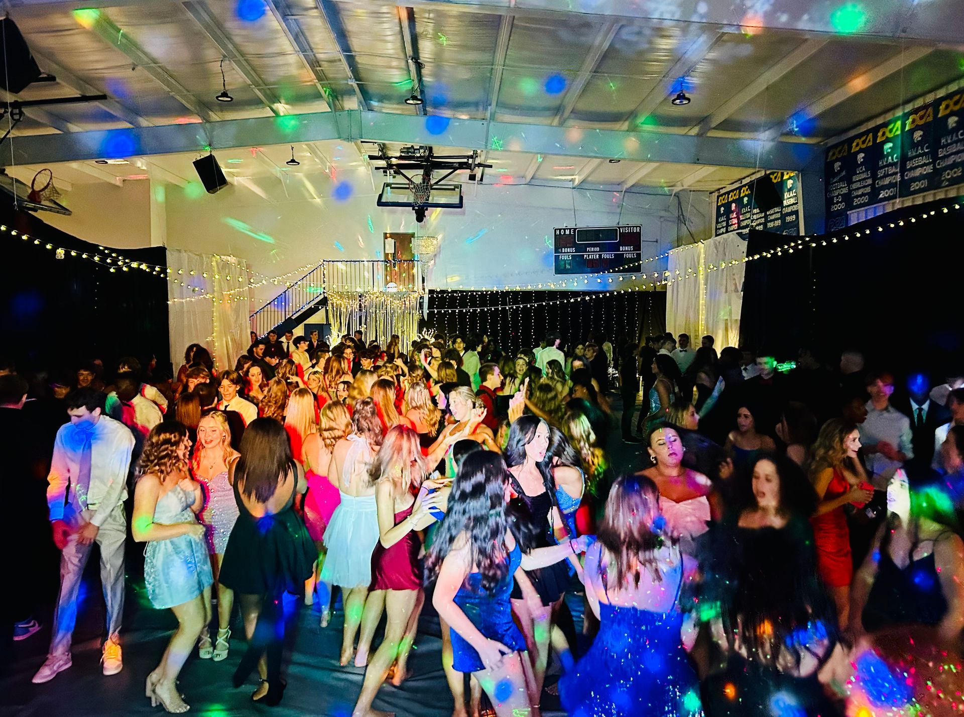 People in formal attire dance and socialize at a dimly lit indoor party with festive lighting and a disco ball effect.