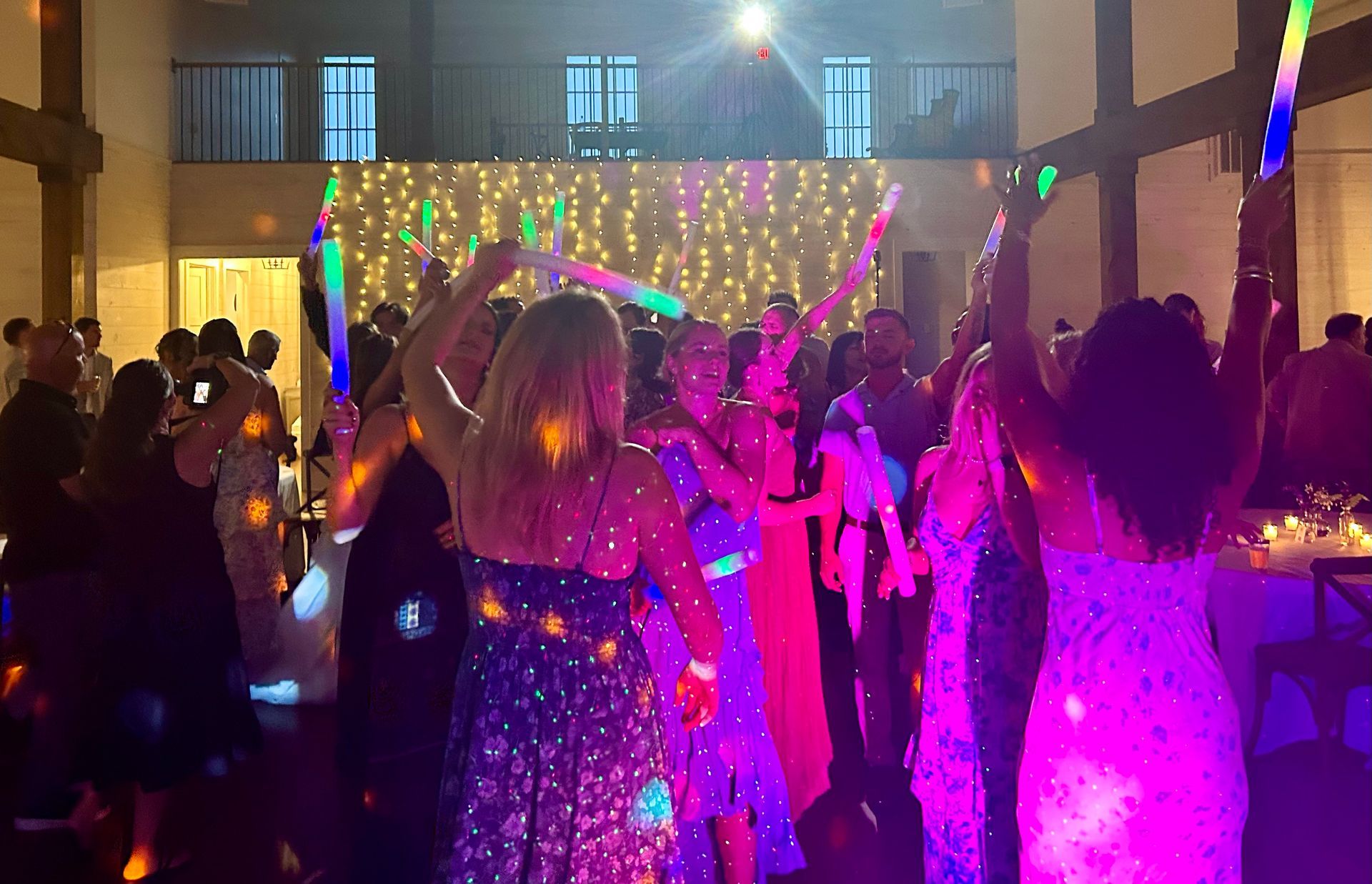 People dancing at an indoor event, holding glowing foam light sticks under vibrant purple and warm string lights.
