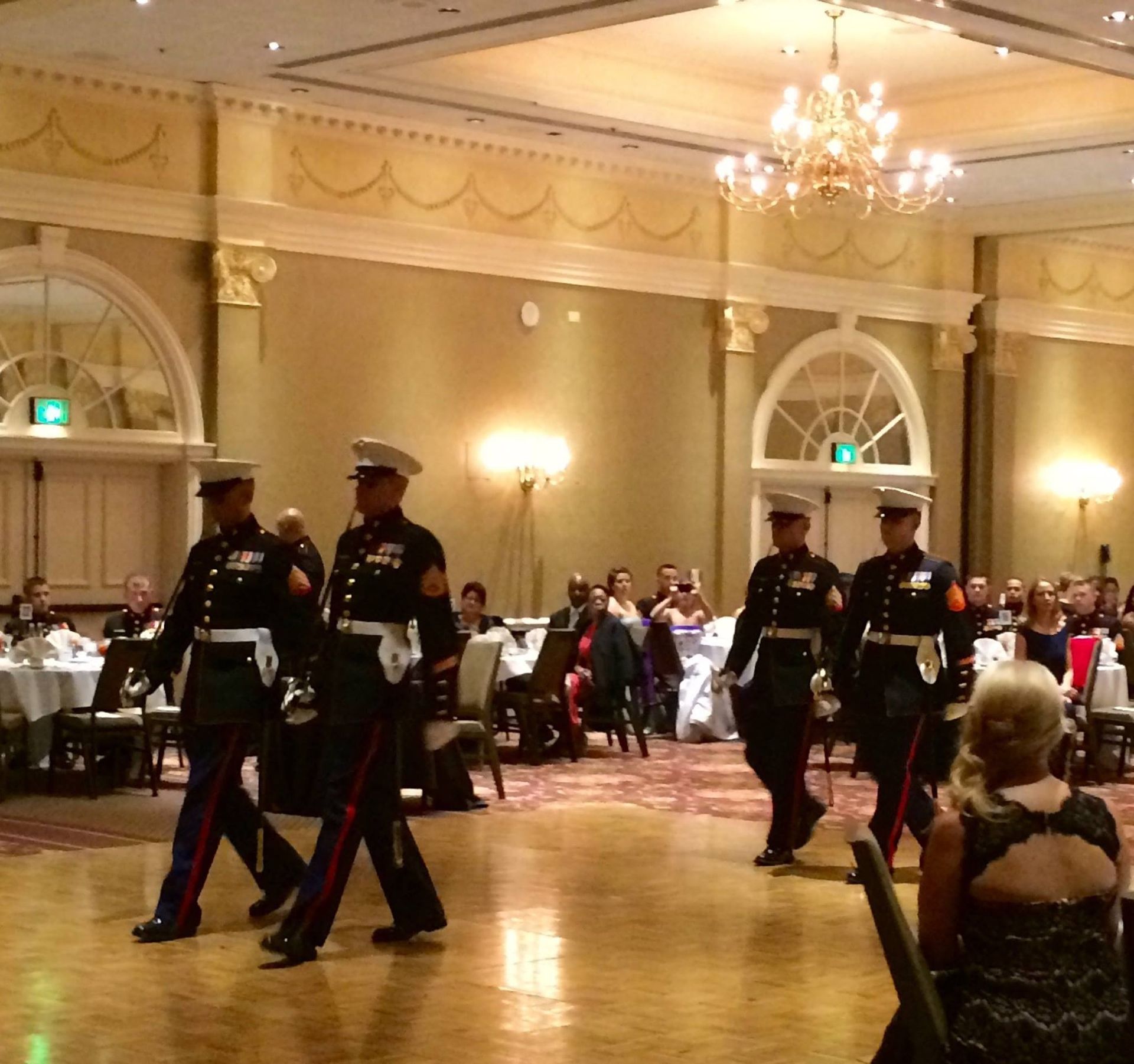 Four uniformed Marines march in a procession across the wooden floor of a formal ballroom event.