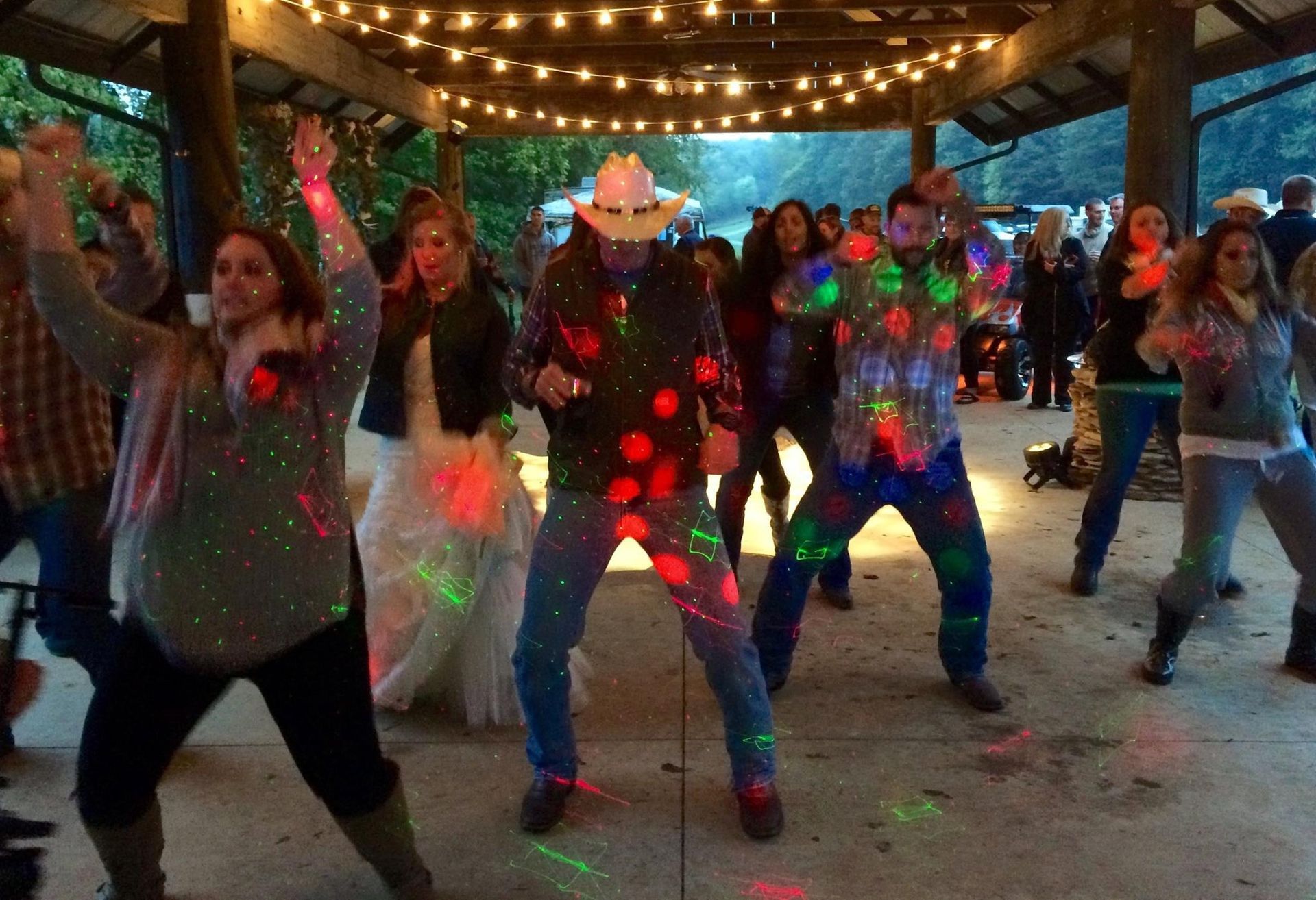 A group of people dancing under string lights at an outdoor event, illuminated by colorful disco light projections.