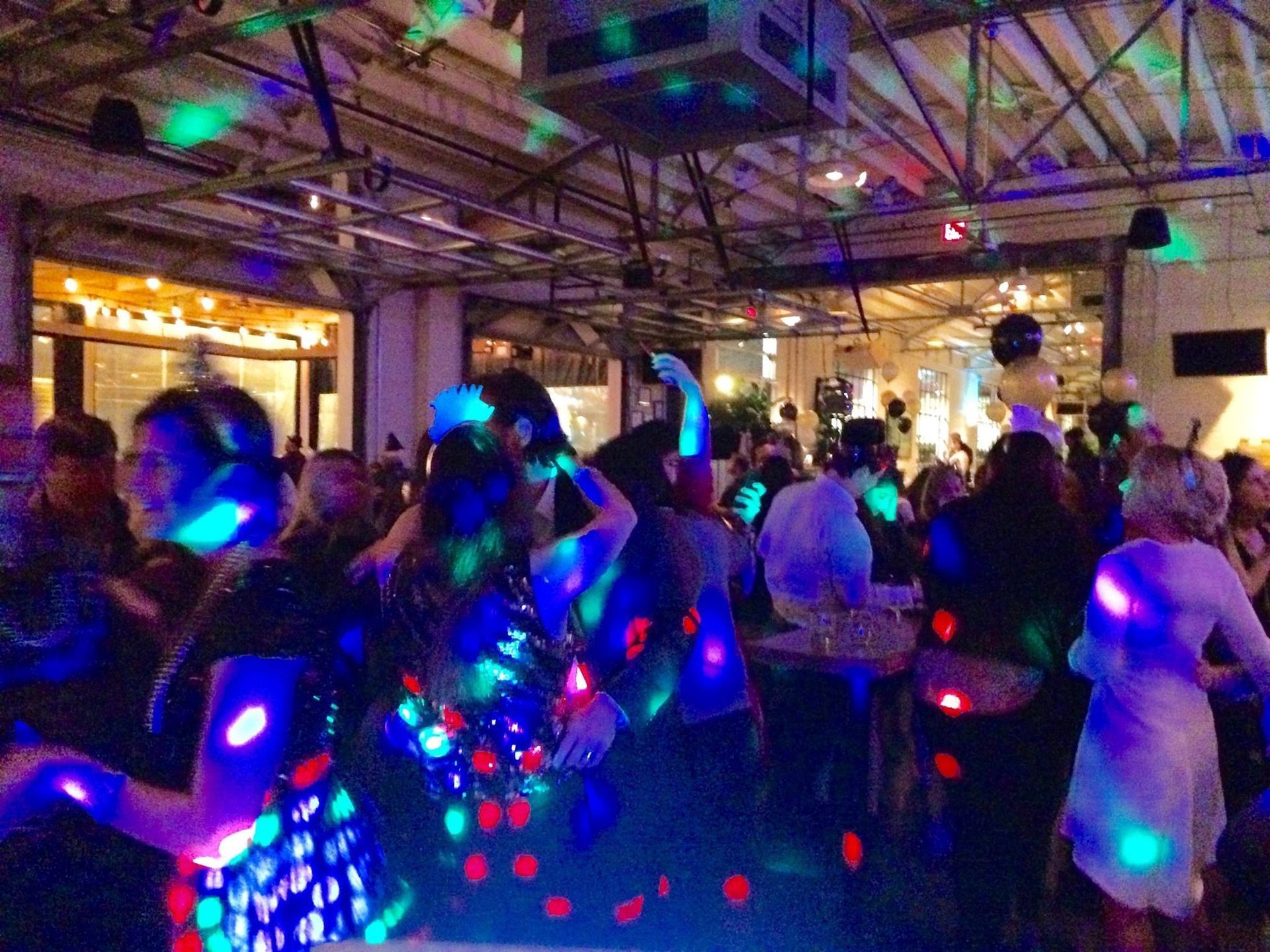 A group of people dancing in a dimly lit, high-ceilinged venue illuminated by colorful, swirling party light effects.