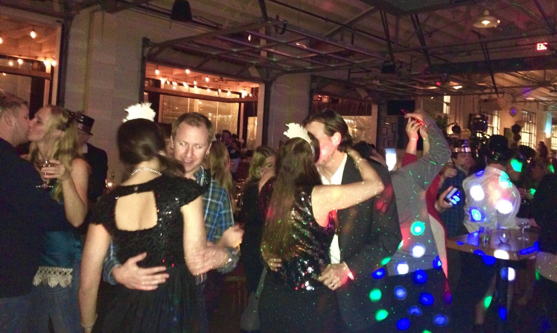 People in formal attire dance and socialize at a dimly lit indoor party with festive lighting and a disco ball effect.