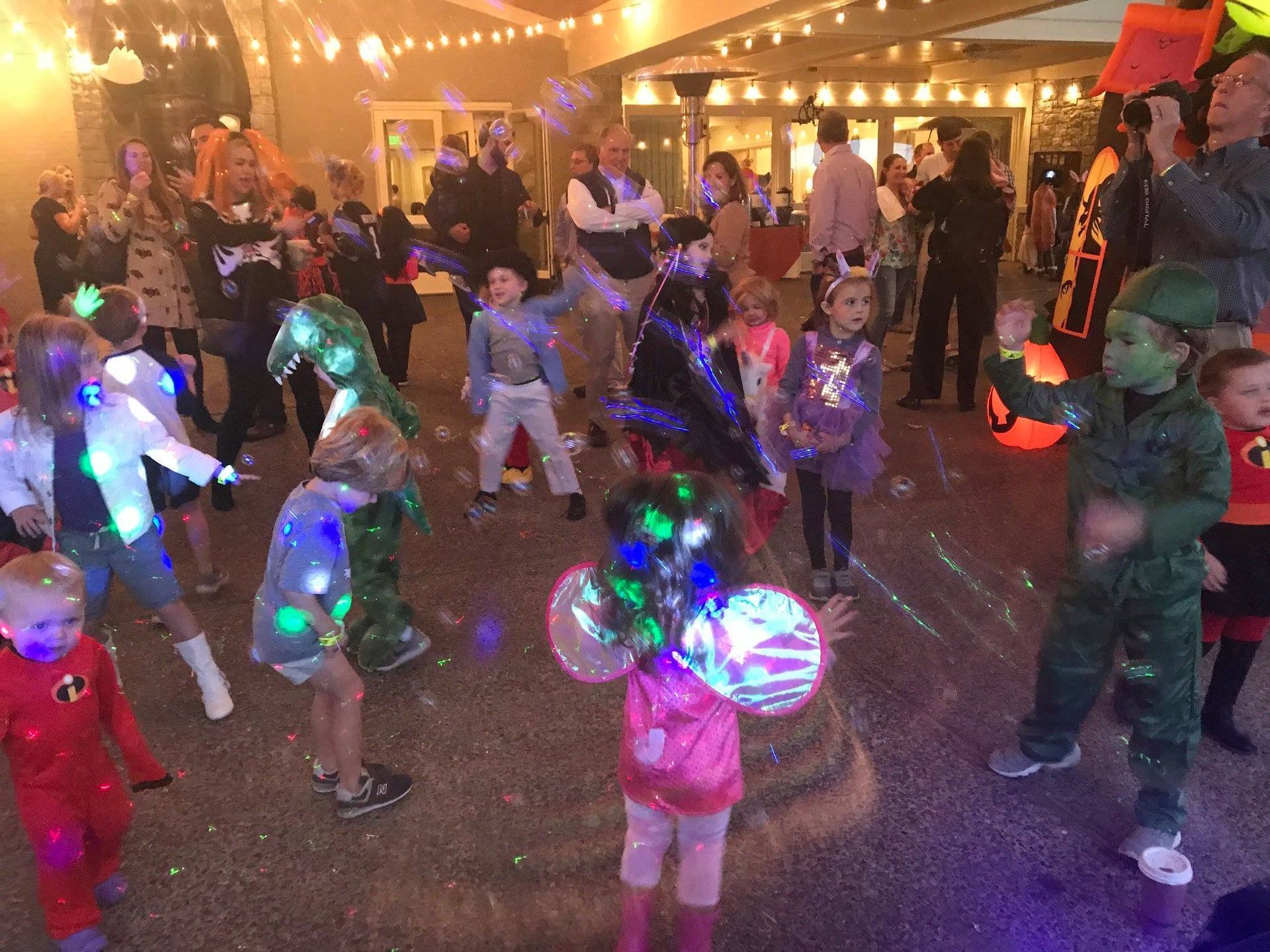 Children in various costumes dance and play amidst bubbles during an outdoor evening event illuminated by string lights.