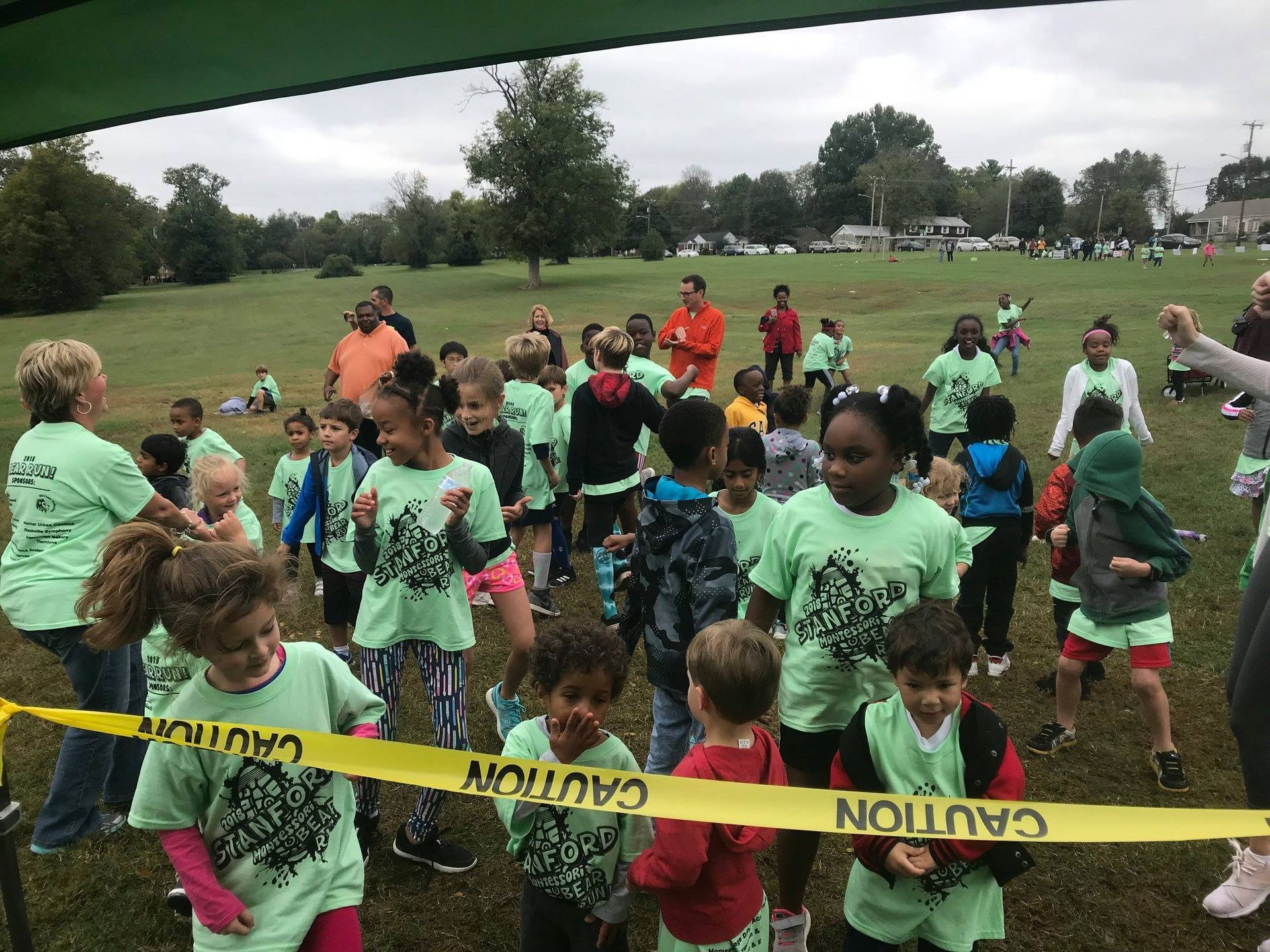 A large group of children and adults in a grassy field, many wearing matching mint green shirts behind a caution tape.