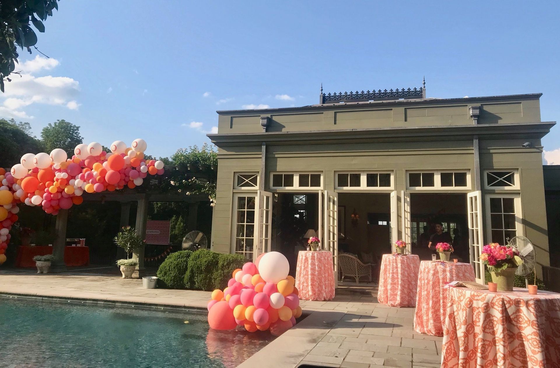 Outdoor event space by a pool decorated with orange, pink, and white balloons and draped tables under a clear blue sky.