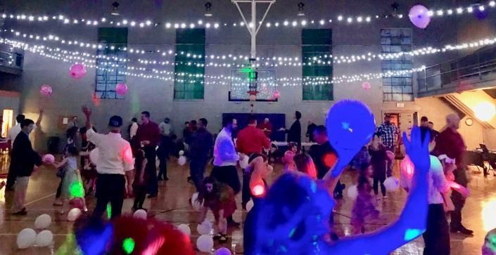 People dance in a gymnasium decorated with string lights and balloons, illuminated by colorful party lighting.