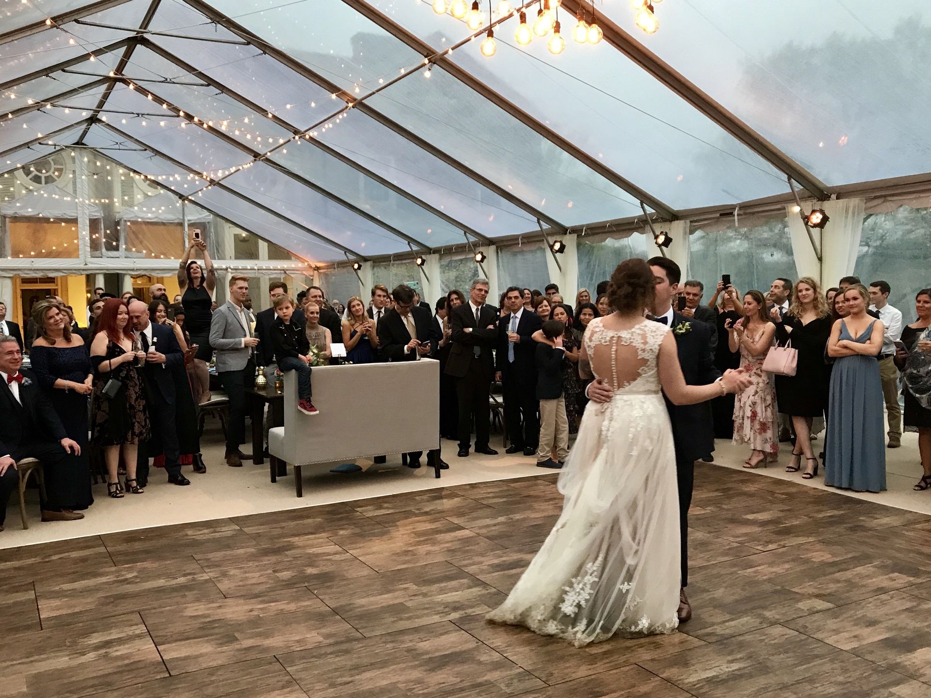 A bride and groom share a first dance on a wooden floor inside a glass-walled event tent decorated with string lights.