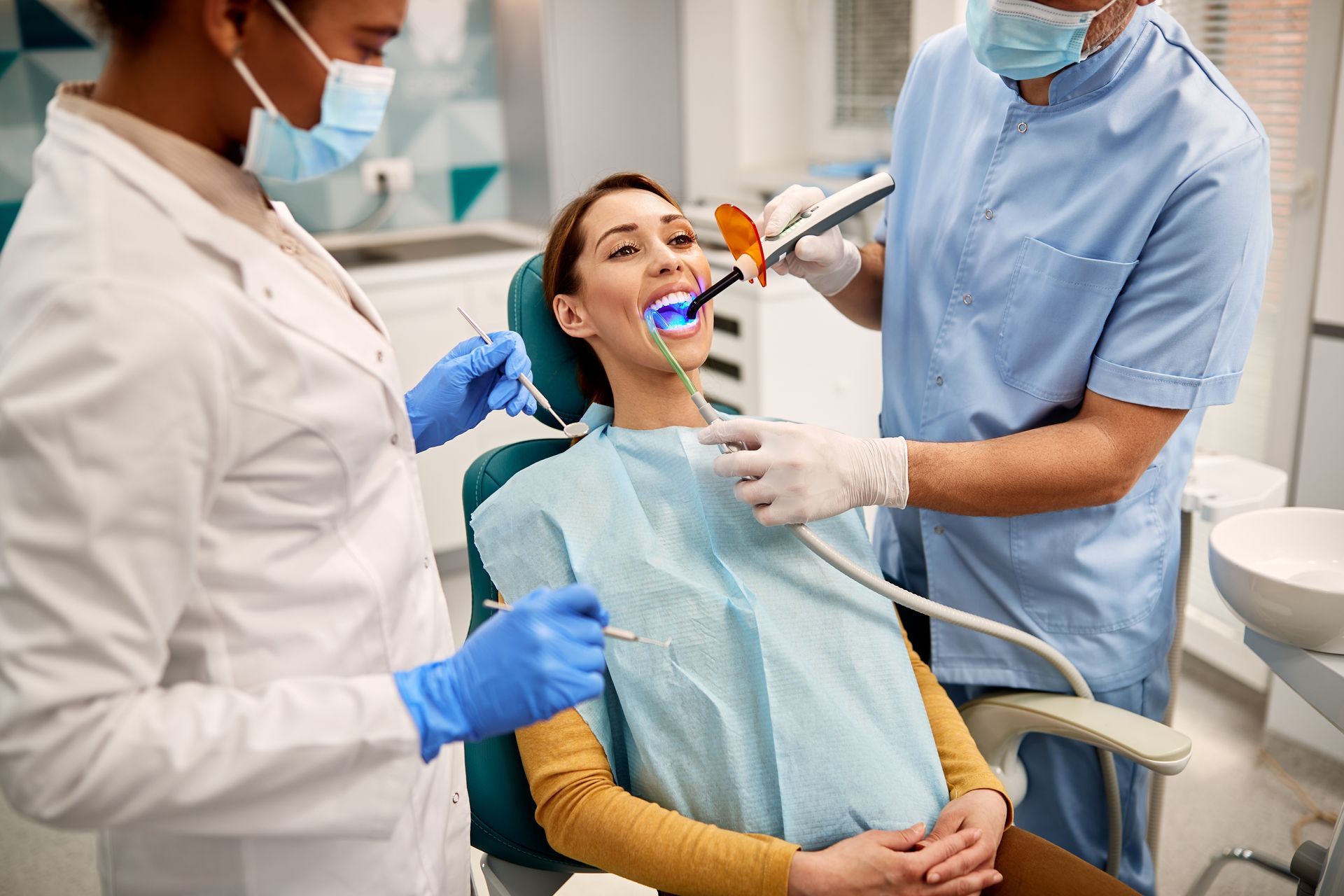 Woman in dentist's chair, being treated by two dental professionals. One uses a curing light.