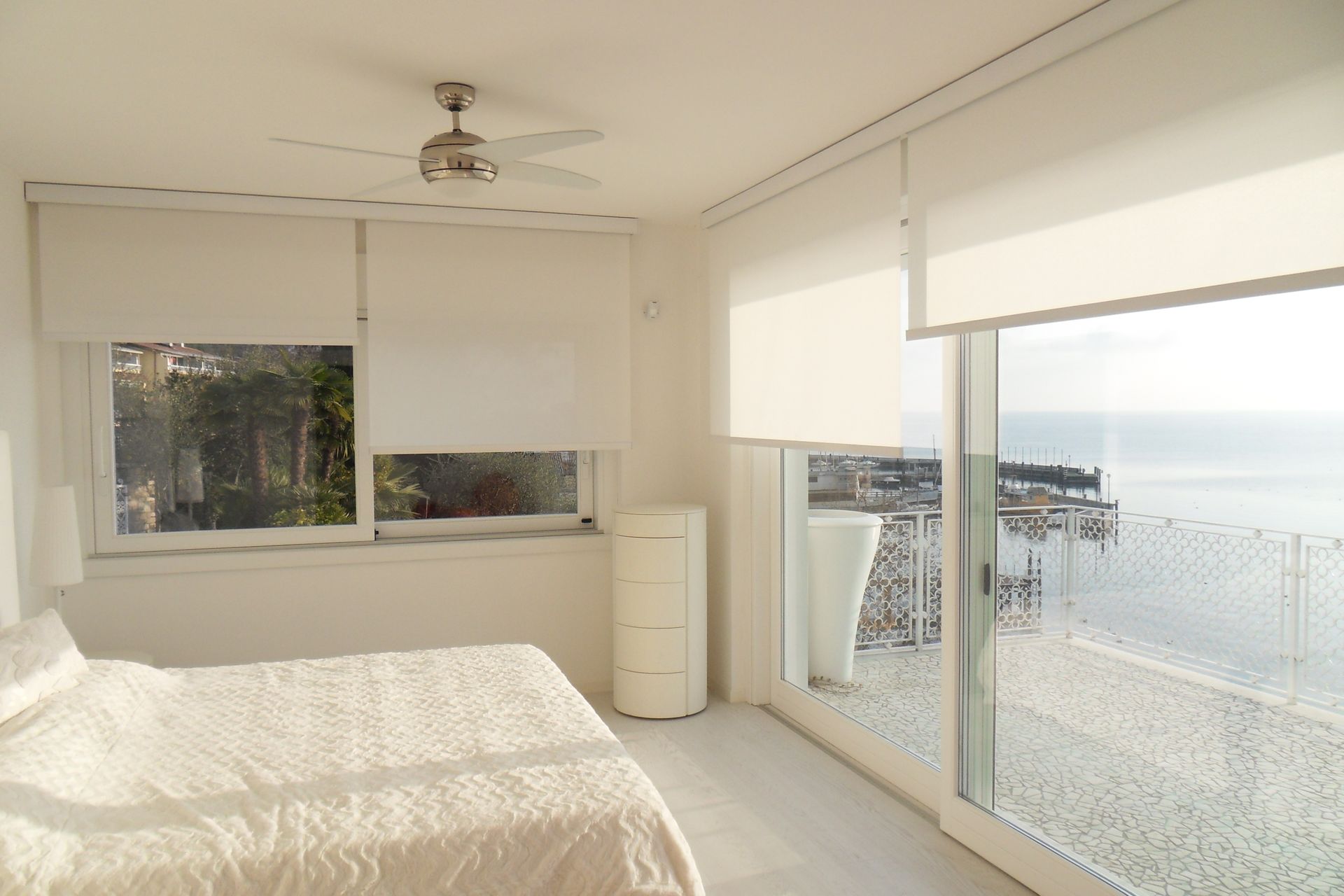 Camera da letto bianca con letto, tende a rullo e vista sul mare.