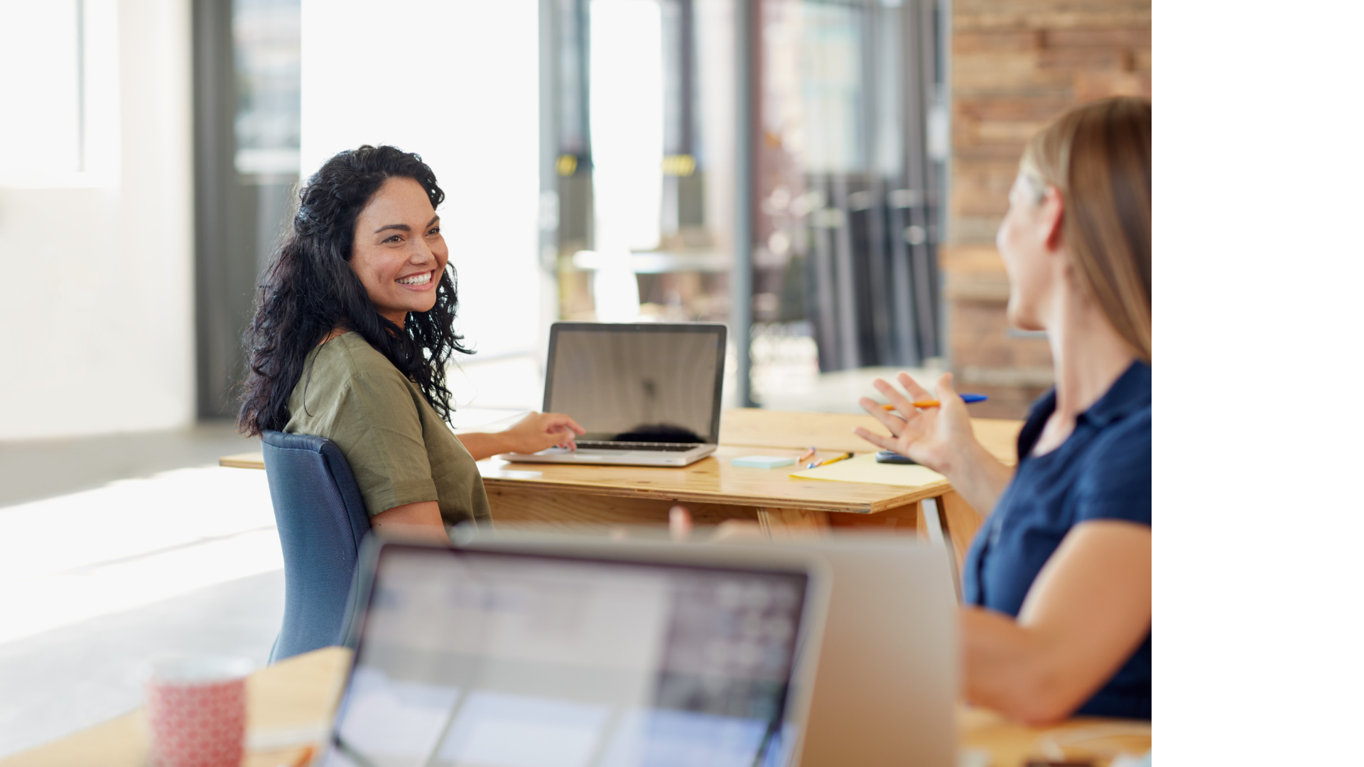 Two women smiling at each other, one near a laptop, in an office setting.