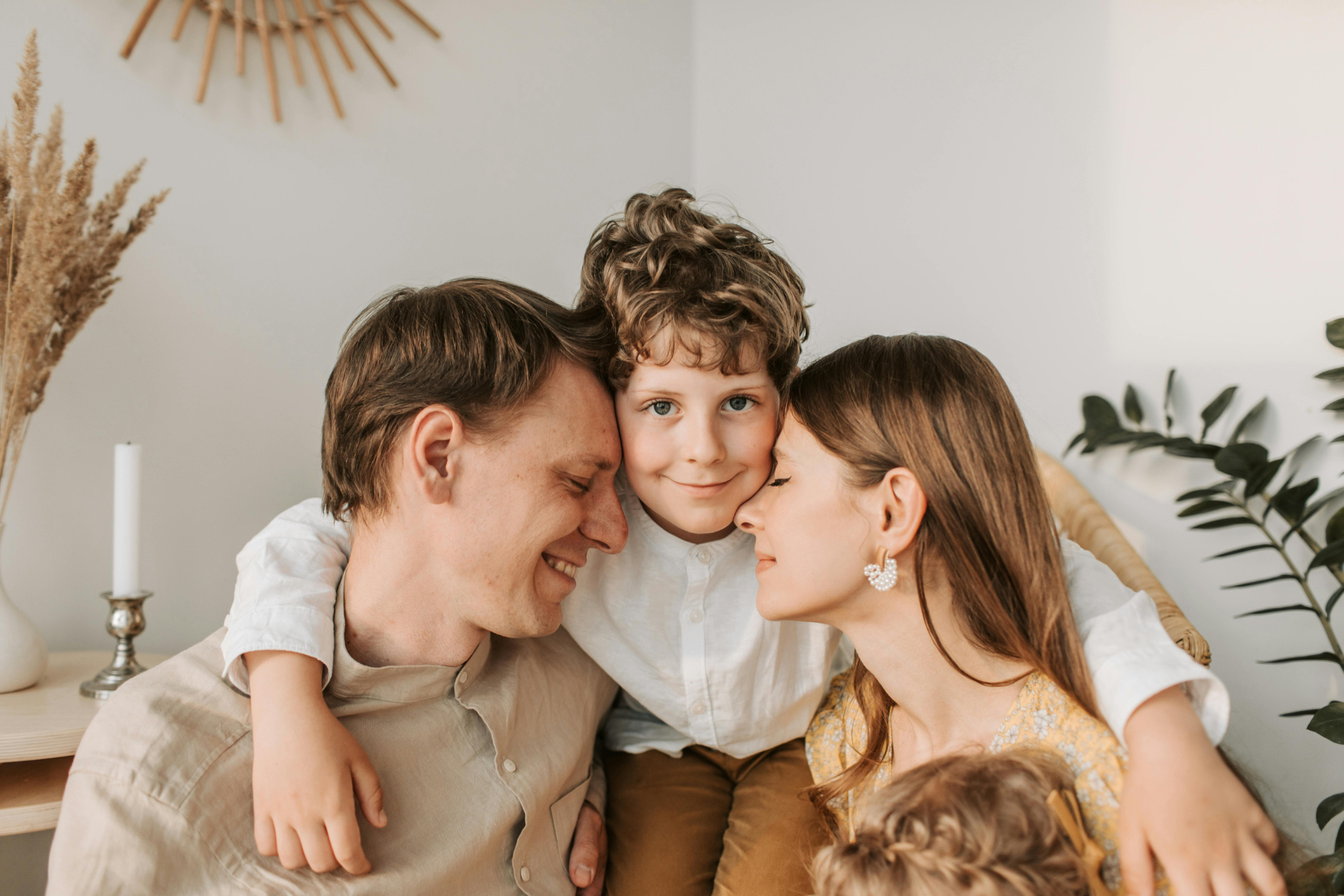 Family of three in a close embrace. Son with curly hair smiles; parents look happy. Interior with plants.