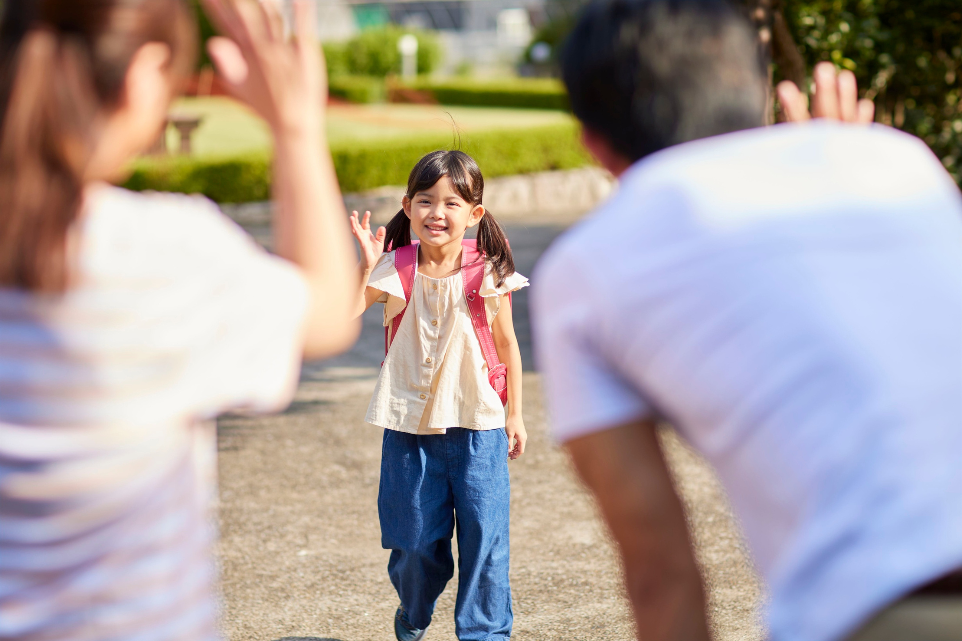 Child with pigtails approaches parents with open arms in sunny outdoor setting.