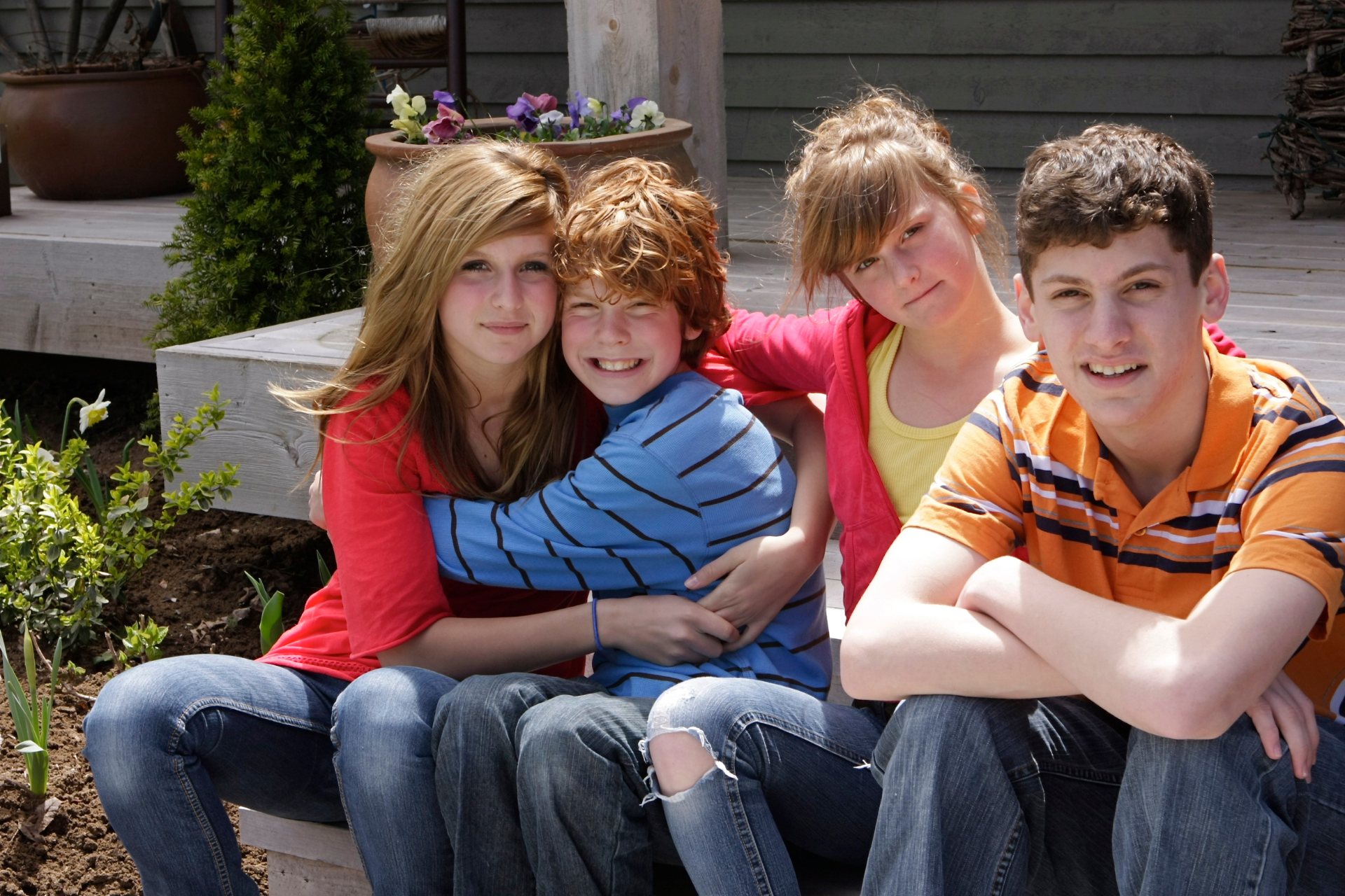 Four children sit on outdoor steps. One hugs another, two look at the camera. Bright colors and sunny setting.