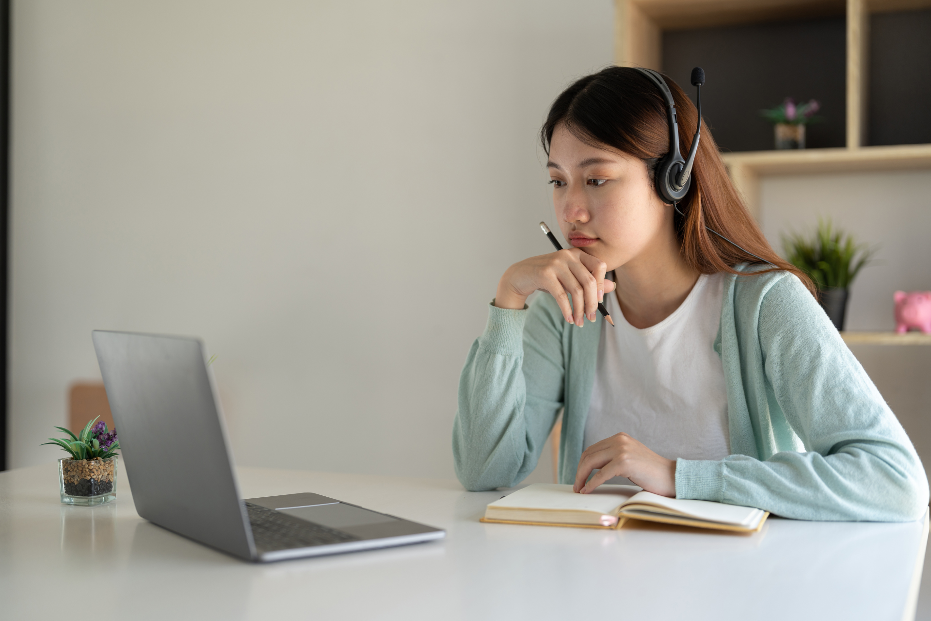 Woman wearing headset, attending a video call on a laptop, taking notes in a notebook.