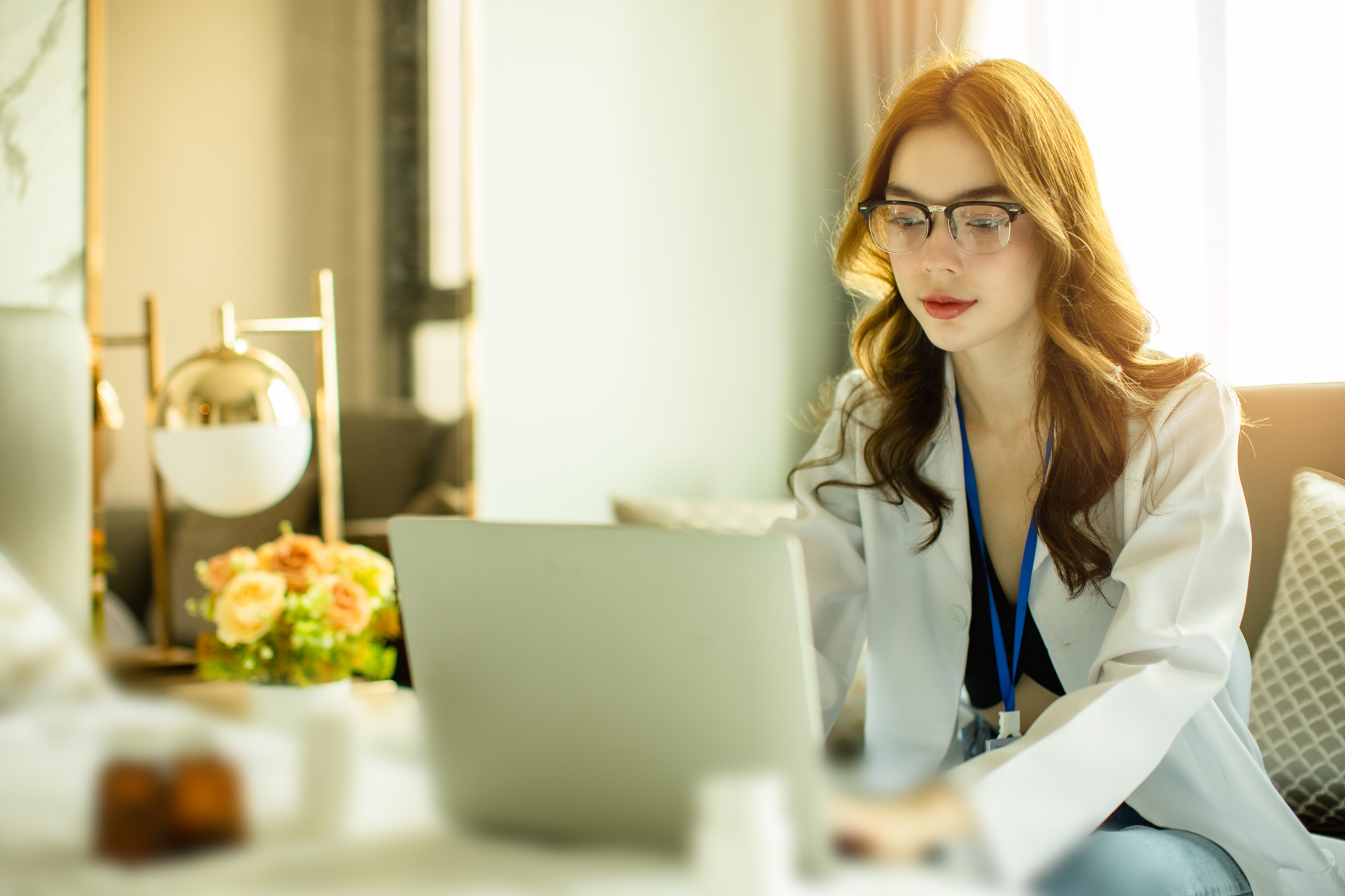 Woman in glasses and white coat using a laptop in a well-lit living room.