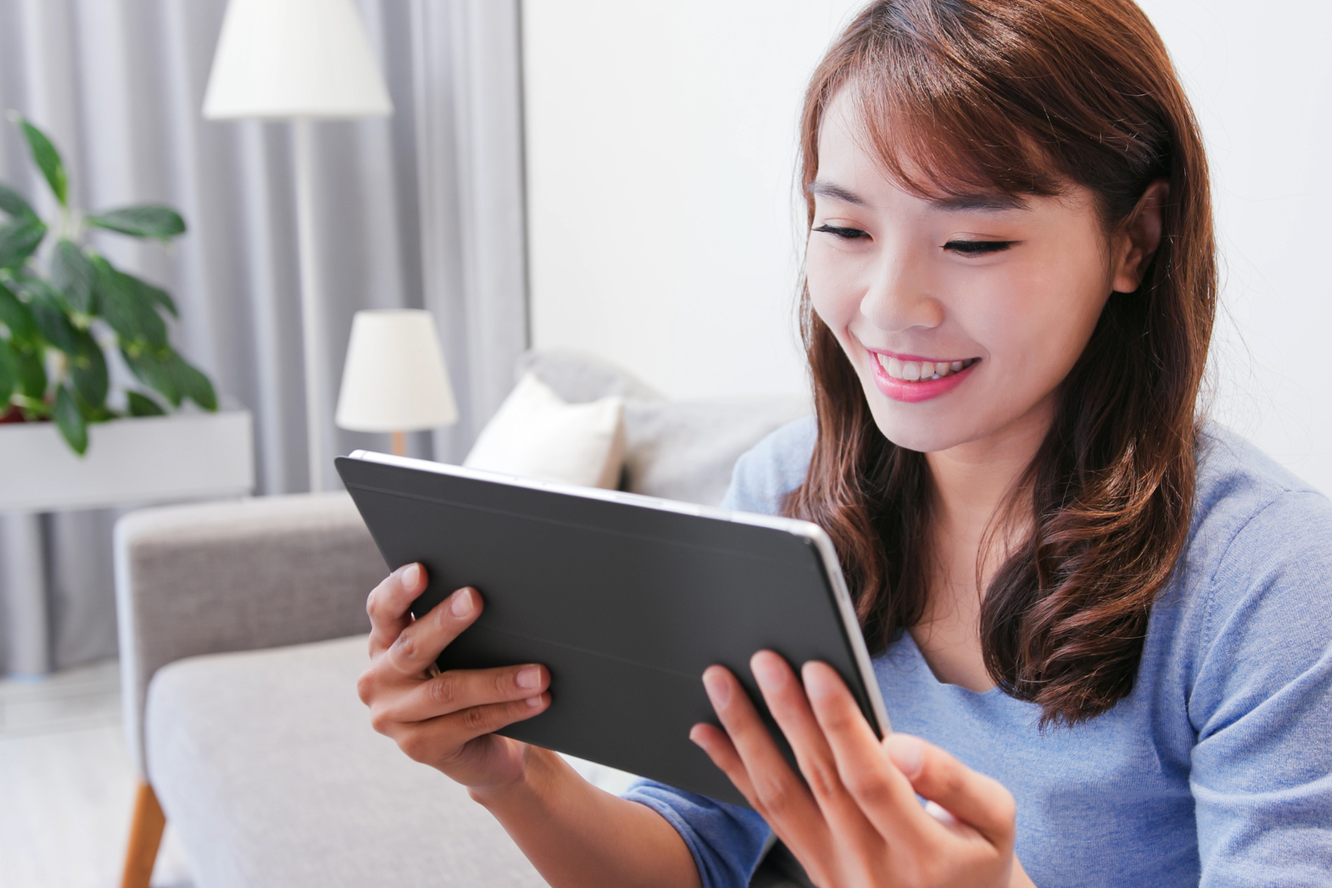 Woman smiling, holding a tablet, sitting on a sofa in a living room.