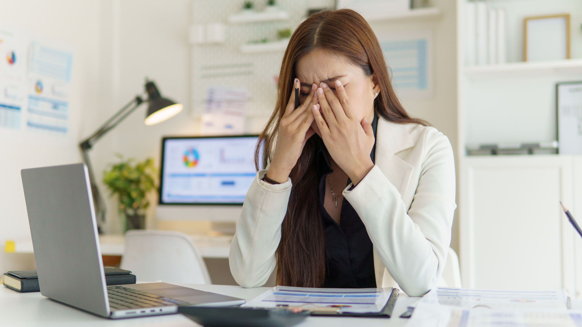 Woman in white blazer, rubbing eyes at a desk with a laptop, monitor, and paperwork.