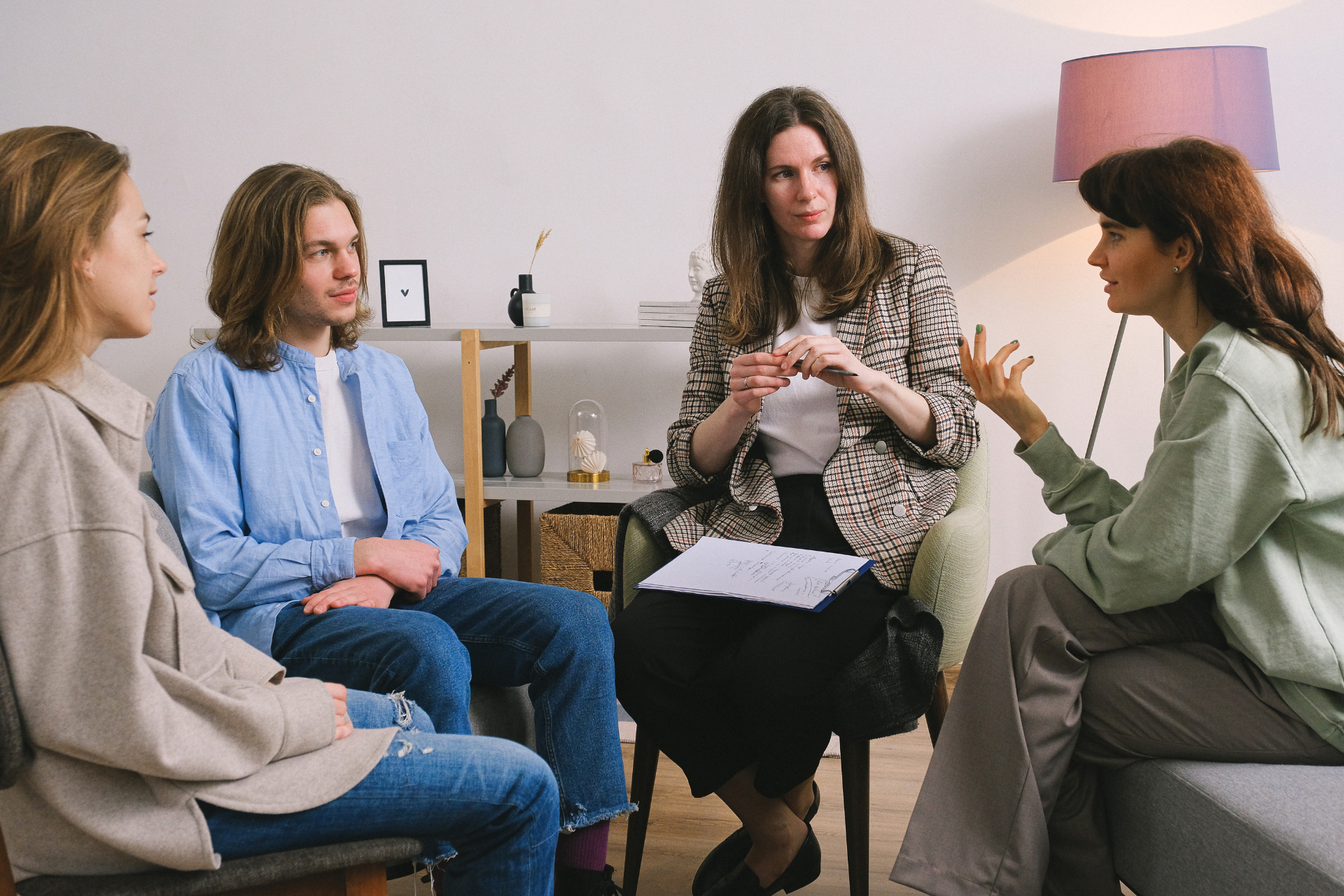 Four people in a circle, one leading a discussion with gestures. Neutral, brightly lit room with furniture.