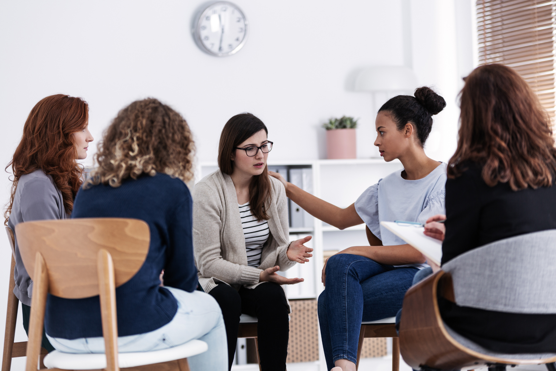 Group therapy session: women in a circle, one woman comforts another, soft lighting.