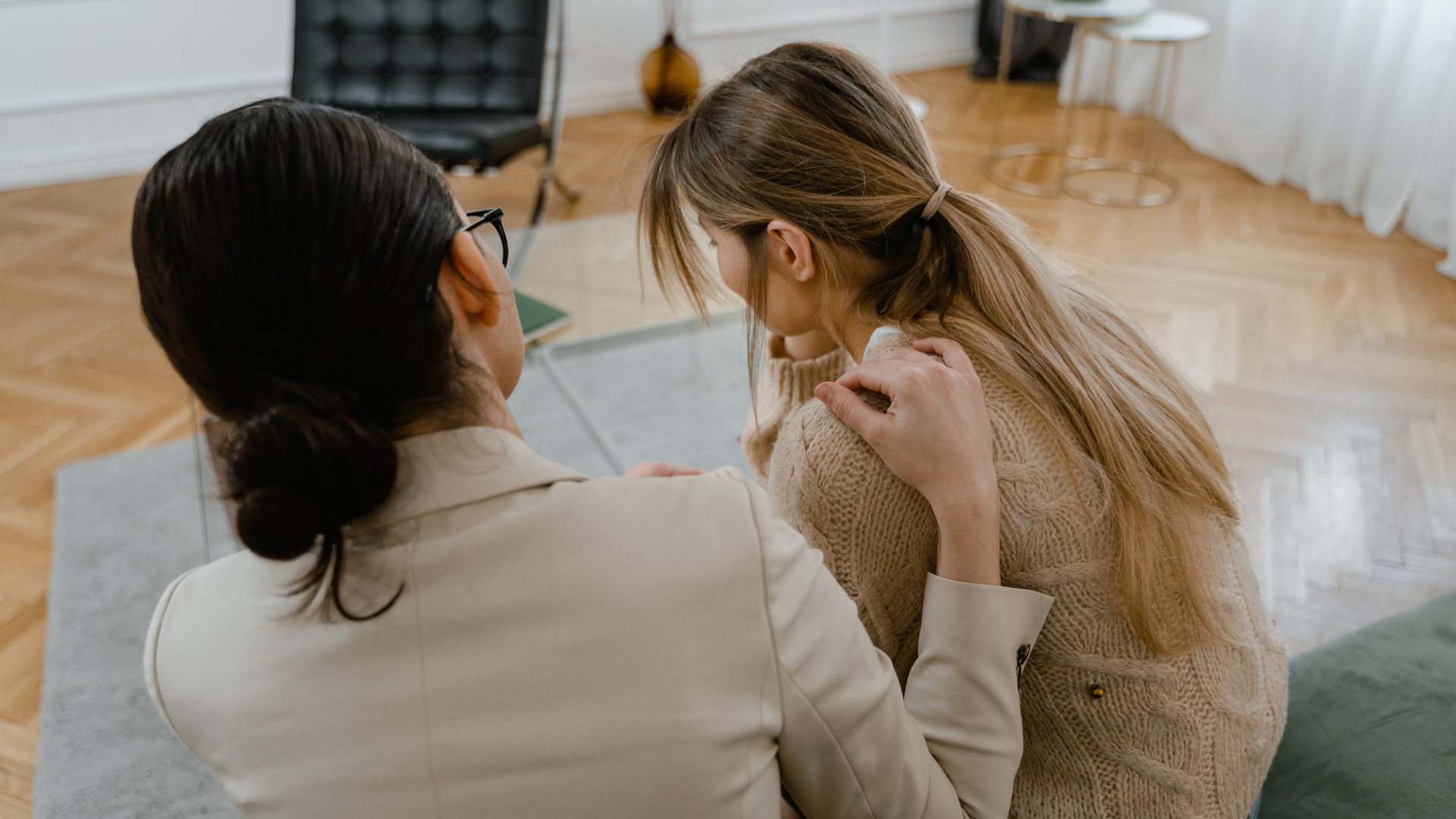 Woman comforting another; one hand on her shoulder, indoors.