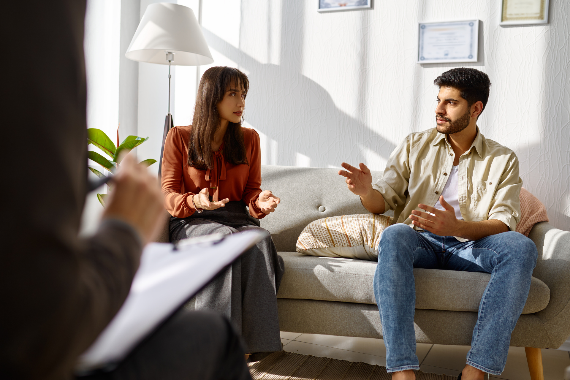 Couple sitting on a couch, talking with a therapist in a brightly lit room.