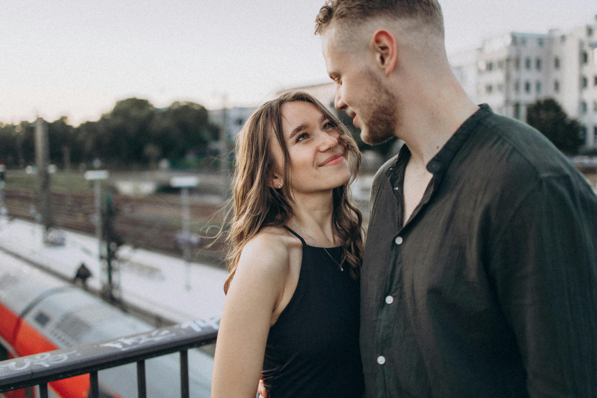 Couple gazing at each other on a bridge with a train passing by. They are close, with soft smiles.