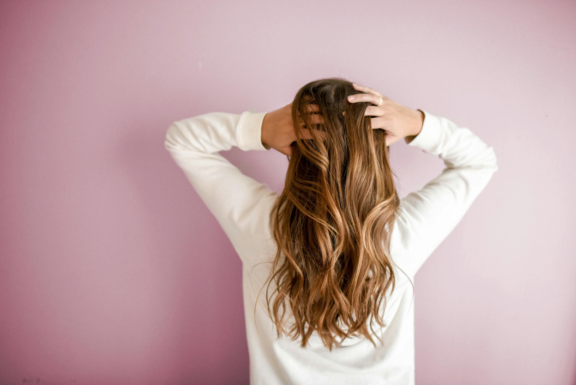 Mujer con cabello castaño ondulado, con las manos en la cabeza, parada frente a una pared rosa.