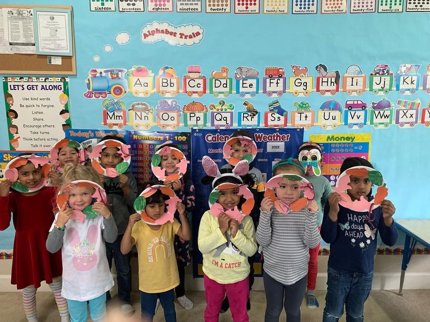 Children standing in front of an alphabet classroom wall, wearing handmade paper hats and holding up their hands.