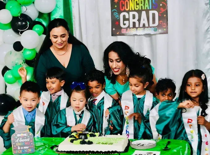 Children and two adults gather around a cake at a graduation party with green-and-black decorations.