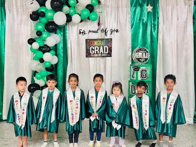 Seven children in teal graduation gowns stand on a decorated stage with black, white, and green balloons.
