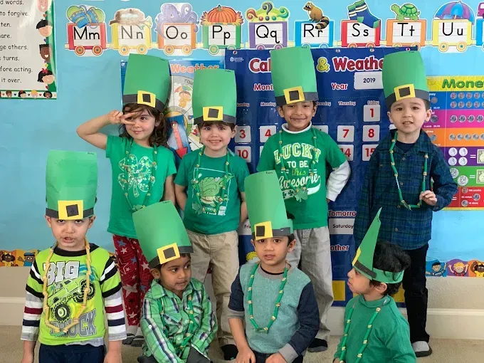 Children in green shirts wearing paper leprechaun hats in a classroom, posing for a group photo