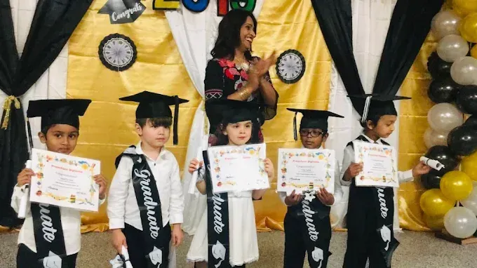 Children in graduation caps and gowns holding certificates on a decorated stage, with an adult behind them