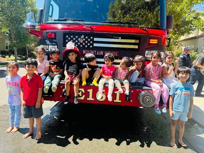 Children posing in front of a red fire truck on a sunny street, some holding hands and smiling