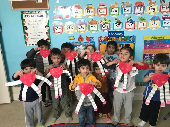 Children in a classroom holding heart-shaped crafts, standing in front of a colorful alphabet wall