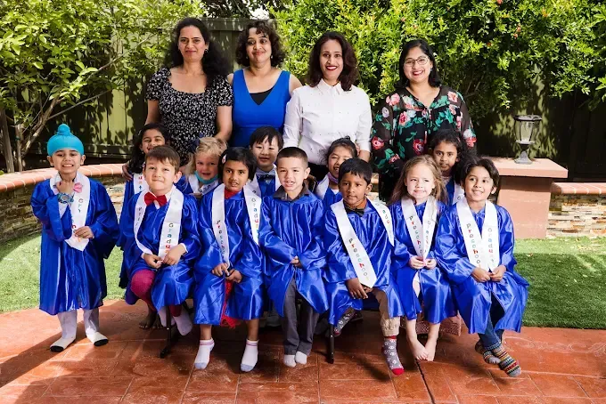 Group of children in blue graduation gowns with four adults standing behind them outdoors