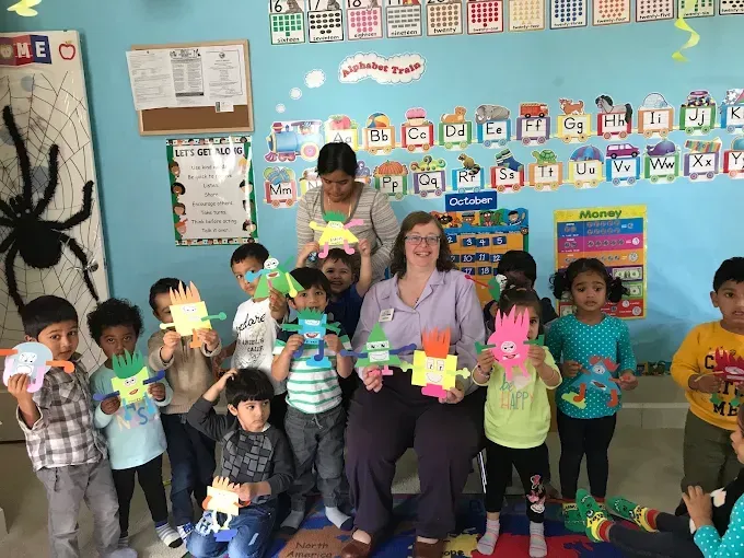 Teacher and children in a classroom holding colorful paper crafts in front of a blue bulletin board.