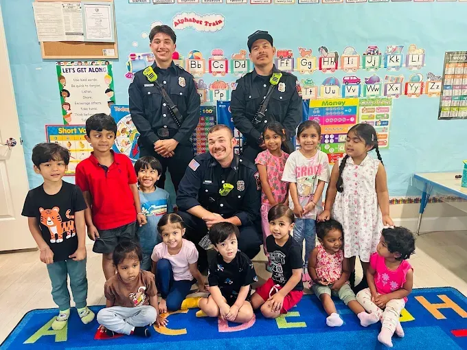 Police officers posing with smiling children in a colorful classroom, seated and standing on a rug.