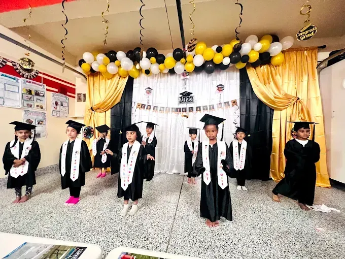 Children in black graduation gowns and caps on a decorated stage with gold and black balloons