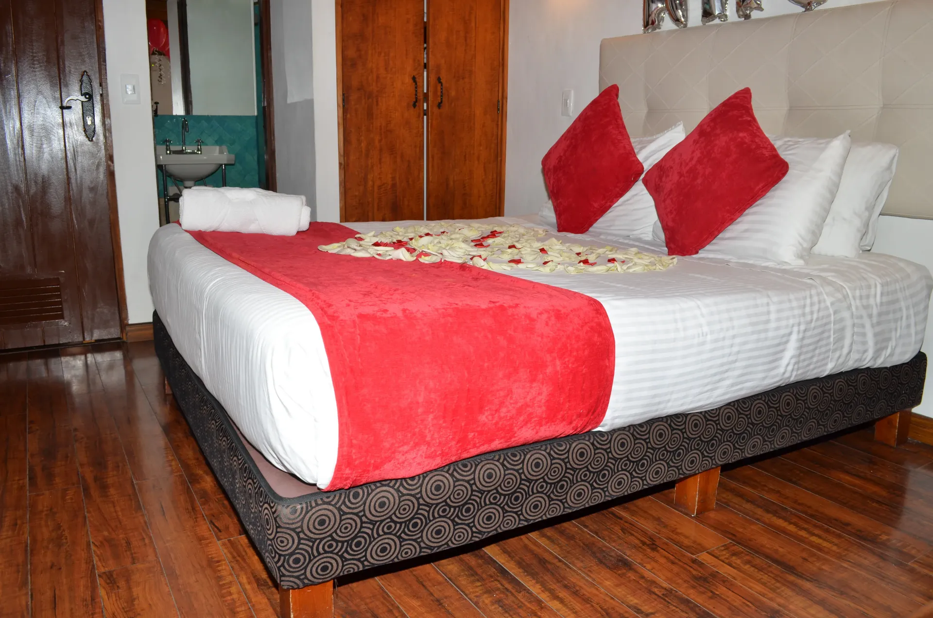 A hotel room bed with red accents and rose petals. Wooden floors and doors are visible.