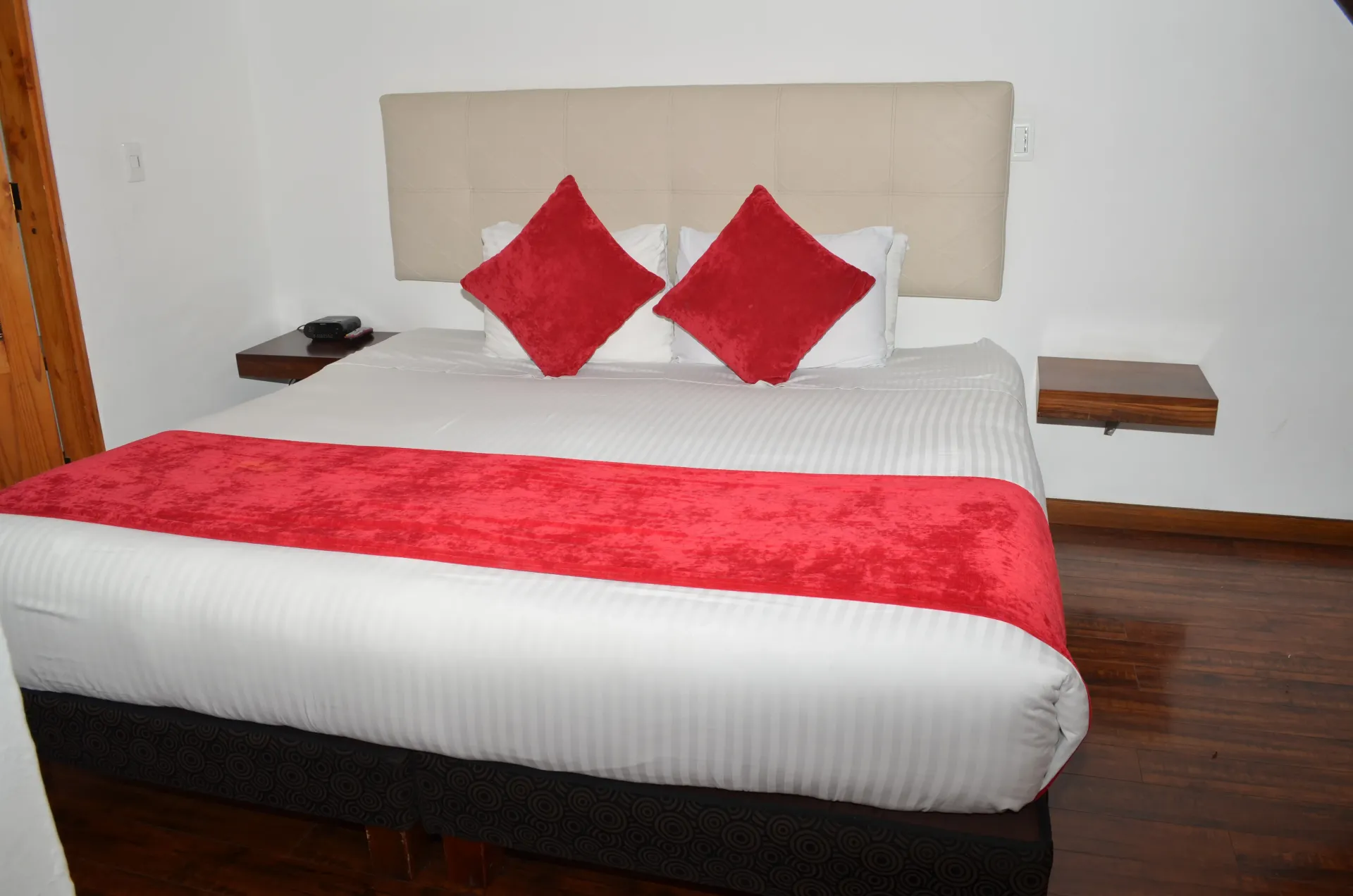 Bedroom with a bed. White linens with red pillows and a runner. Wooden bedside tables and floor.