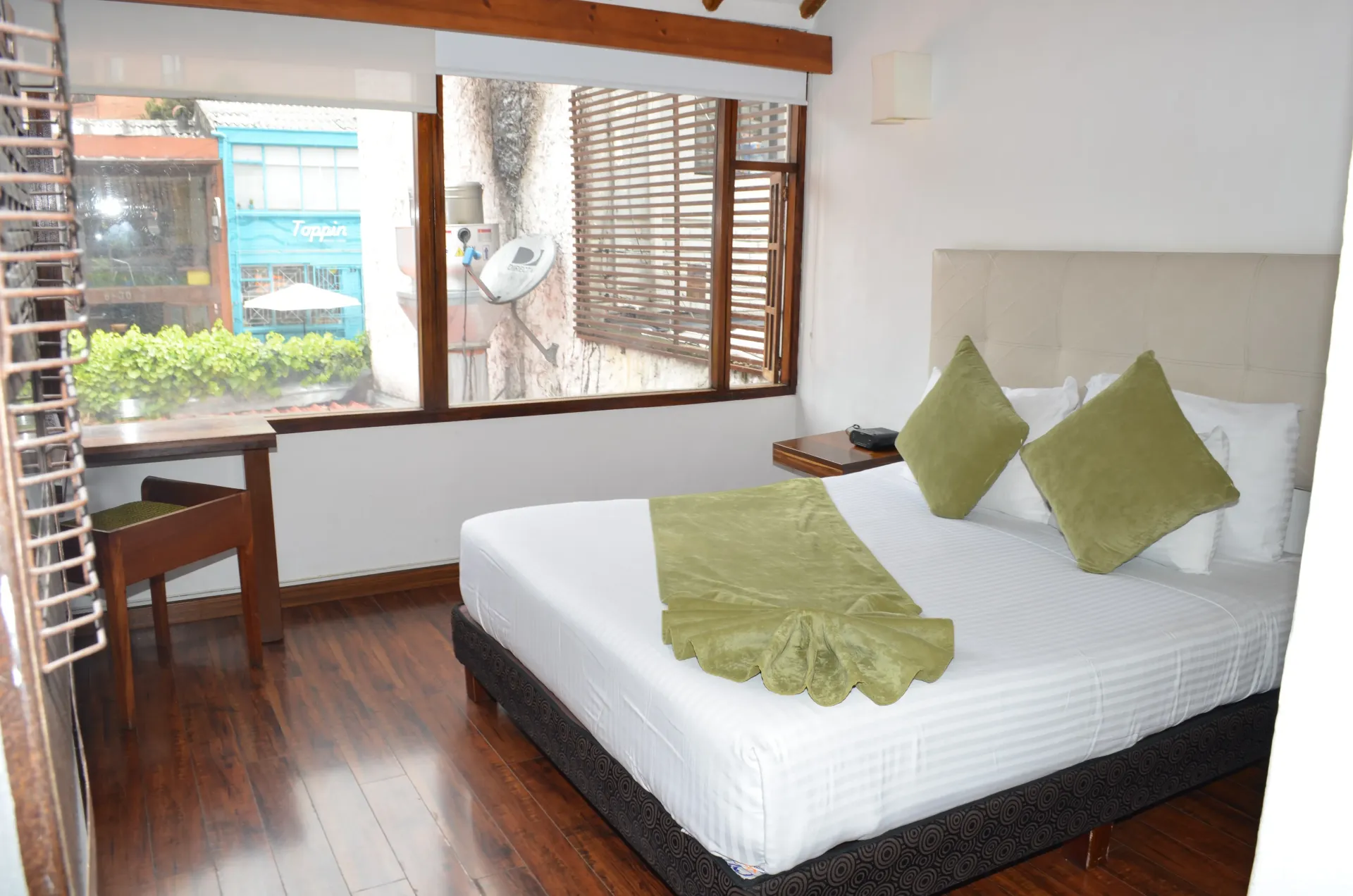 Hotel room with a bed, desk, and window overlooking greenery and a blue building.