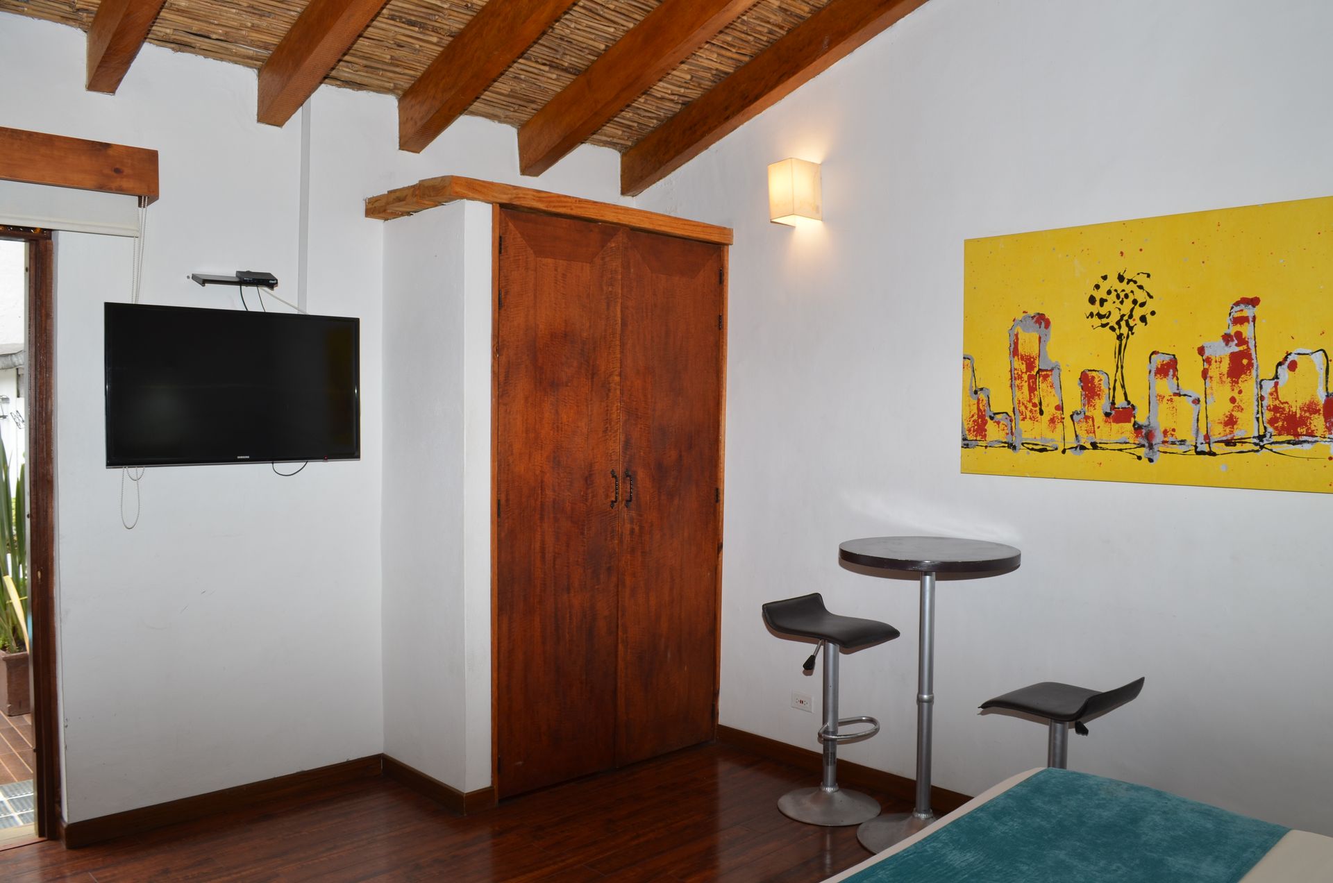 Bedroom with TV, wooden wardrobe, yellow artwork, small table, and stools.
