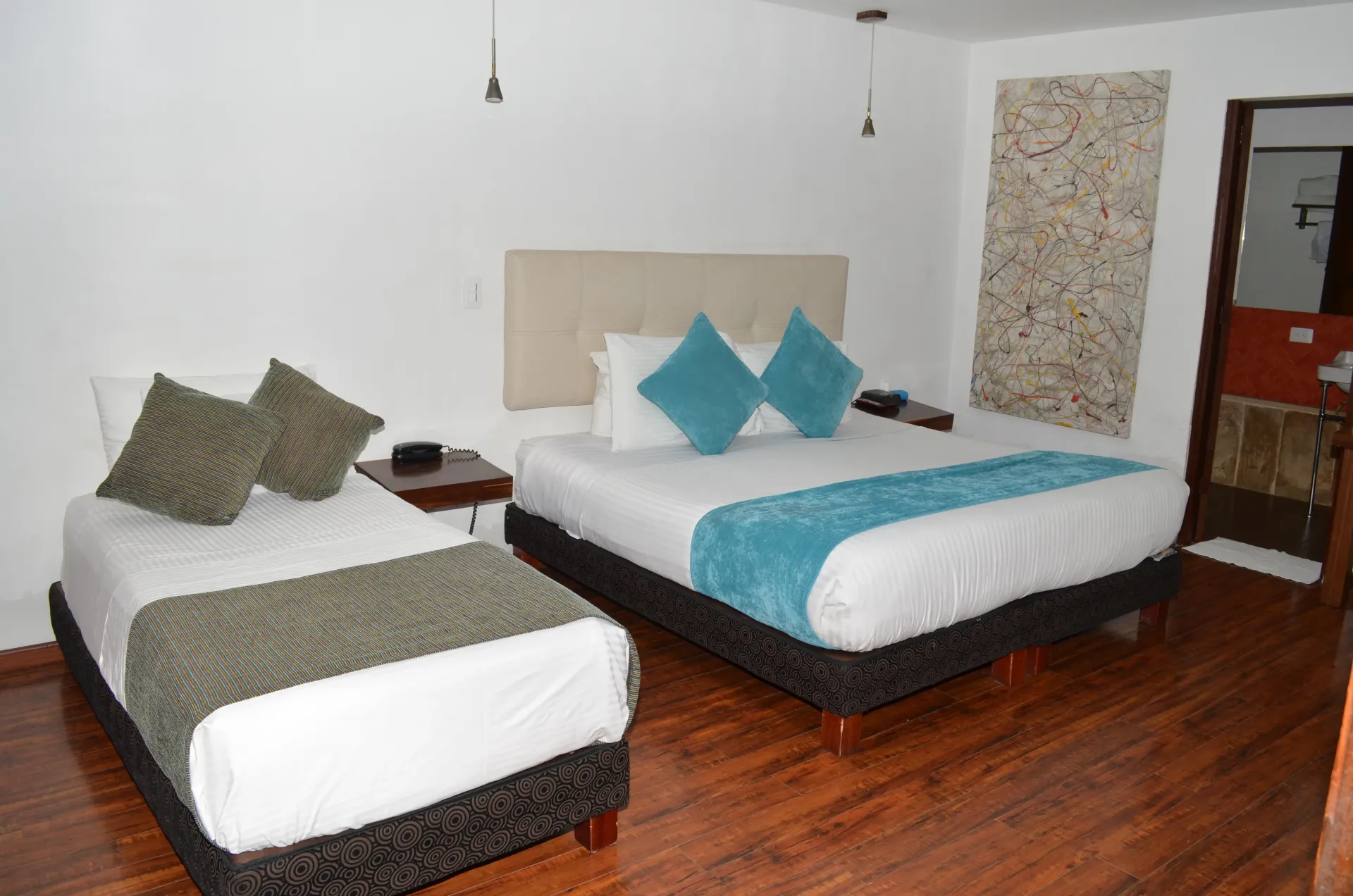 Hotel room with two beds: one queen and one twin, wood floor, white walls, and turquoise and gray accents.