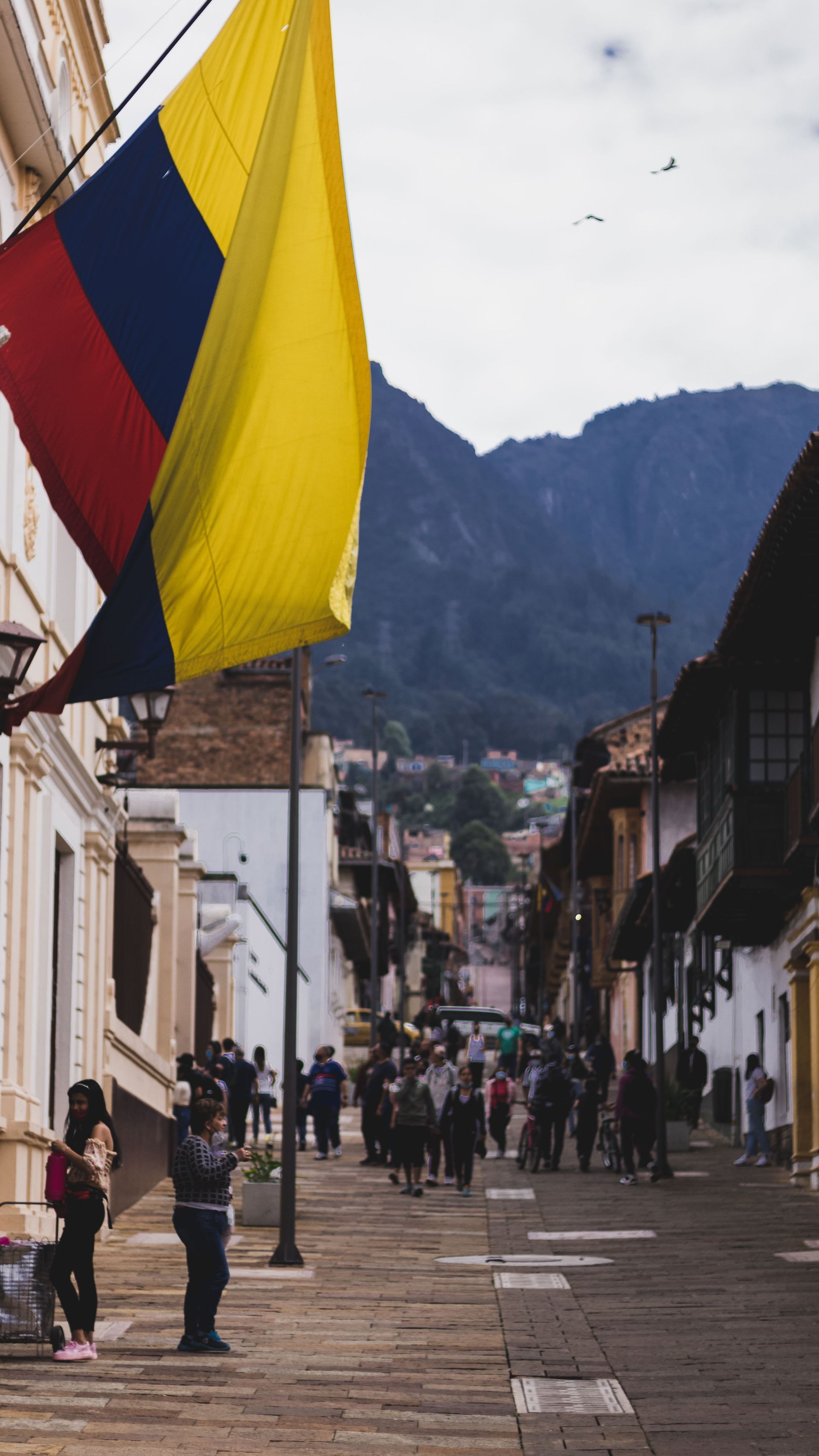 Banderas de colores ondean sobre una calle empedrada por donde camina la gente, con las montañas al fondo.