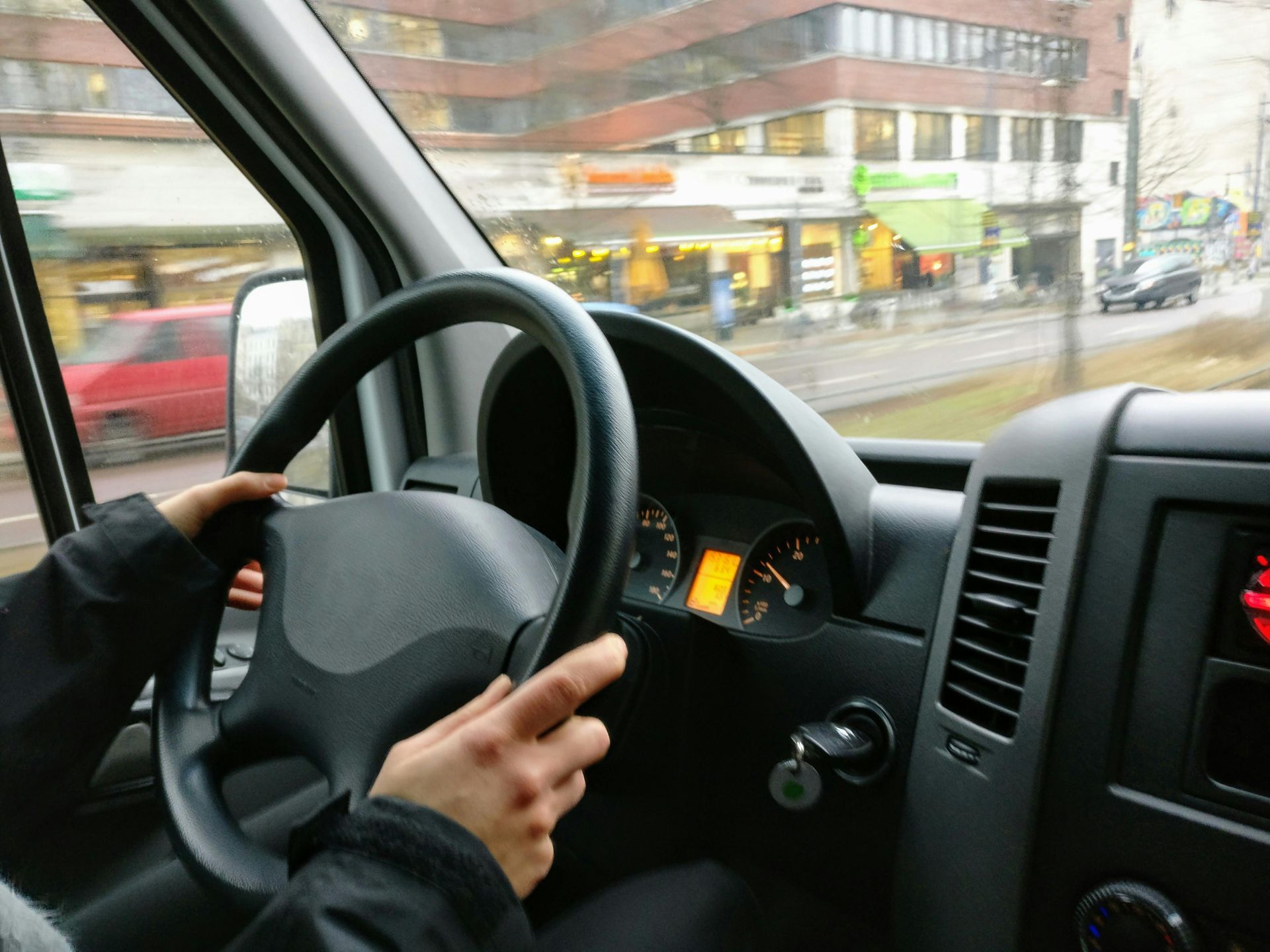 Manos aferradas al volante dentro de una furgoneta, conduciendo junto a edificios en una calle de la ciudad.