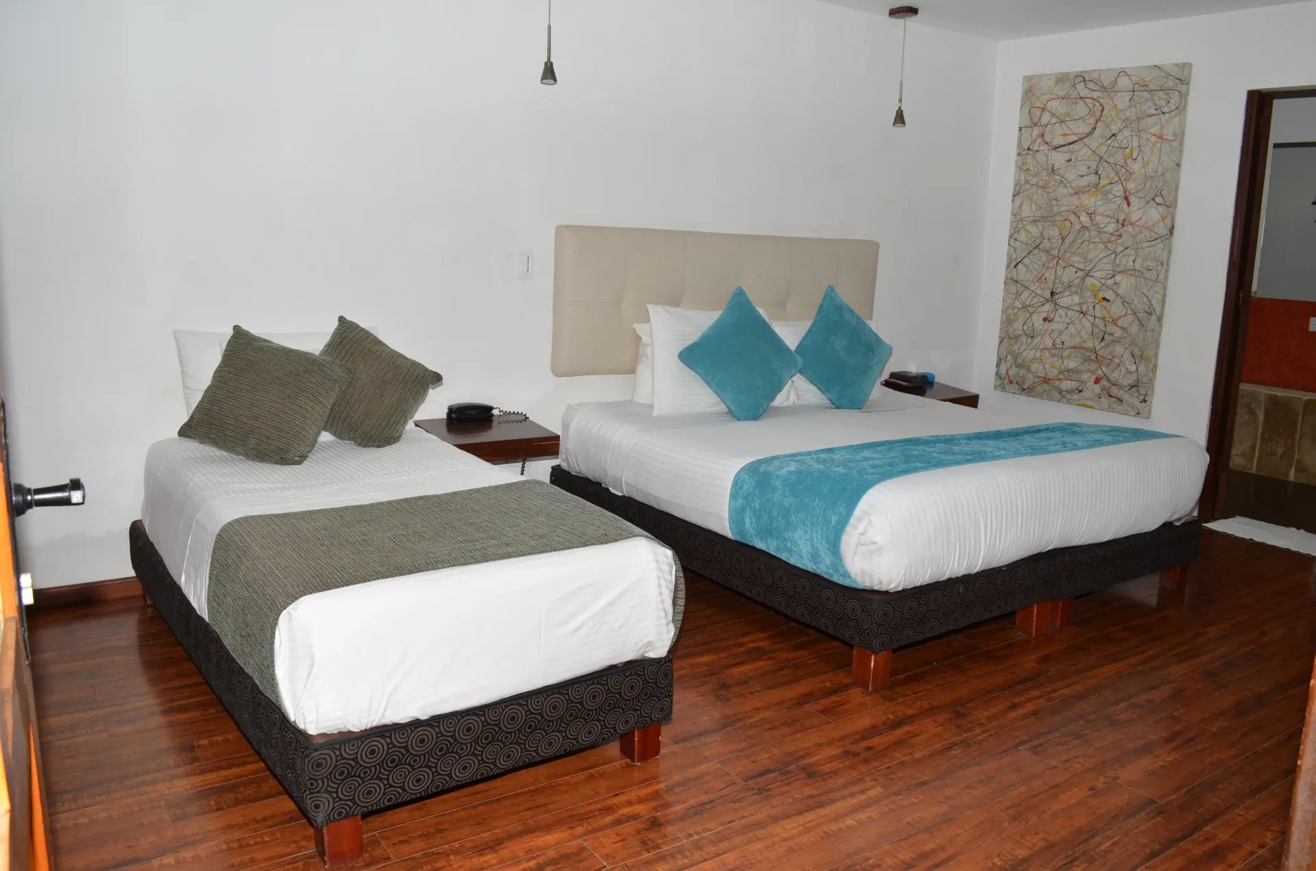 Hotel room with two beds, one queen with teal accents and one twin with green accents, wood floor.