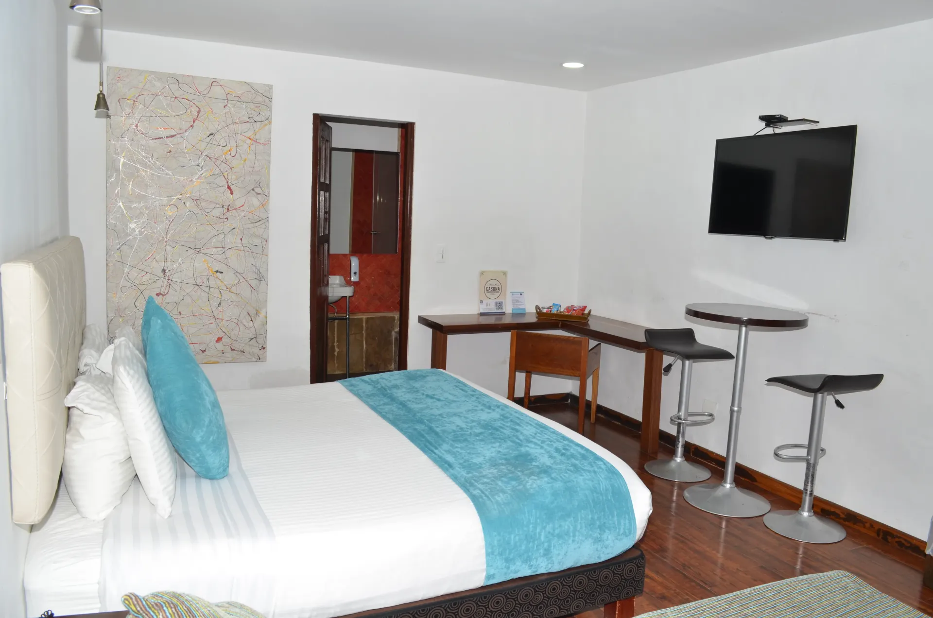 Hotel room with bed, desk, TV, and bar stools. Bright with white walls and wooden floors.
