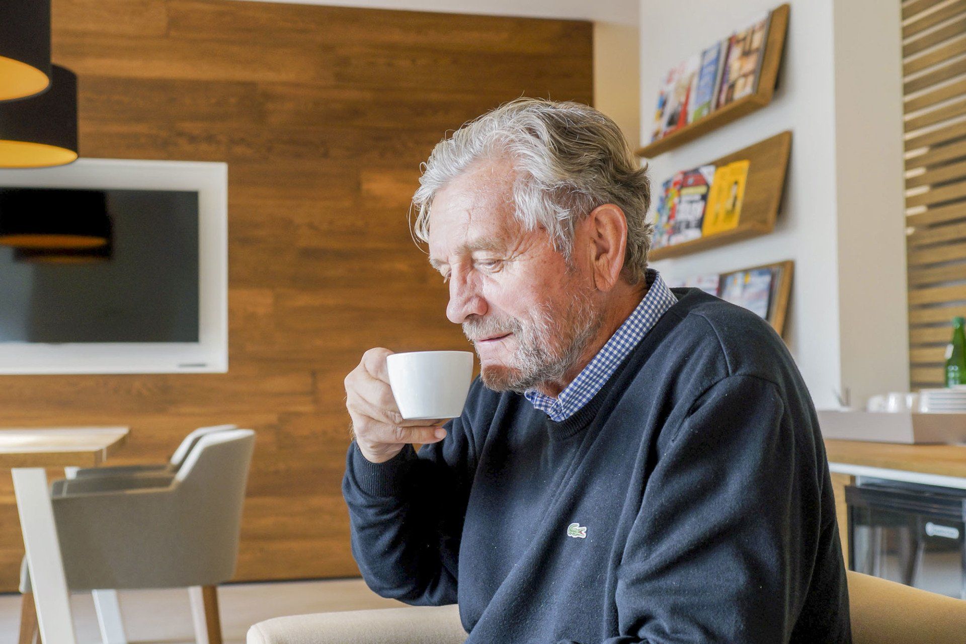 An older man is drinking a cup of coffee while sitting in a chair.