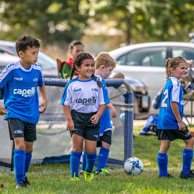 Soccer players in blue and white uniforms on a grassy field, with a soccer ball and cars in the background.