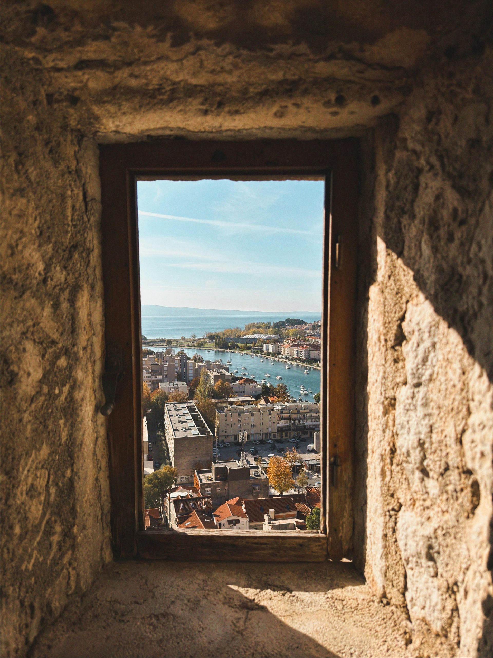 A window in a stone wall with a view of a city and the ocean.